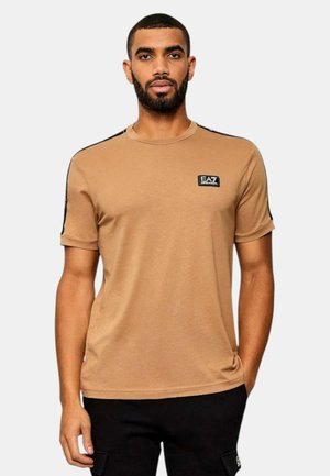 Uomo con barba che indossa una t-shirt color cammello EA7 Emporio Armani e pantaloni neri, in piedi con una mano in tasca contro uno sfondo neutro.