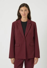 Vrouw met lang donker haar, gekleed in een bordeauxrode blazer en bijpassende broek over een zwarte top, staand tegen een eenvoudige lichte achtergrond.
