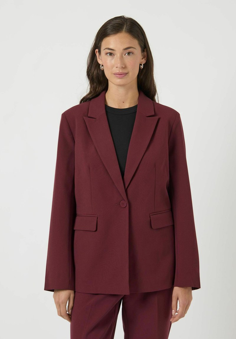 Vrouw met lang donker haar, gekleed in een bordeauxrode blazer en bijpassende broek over een zwarte top, staand tegen een eenvoudige lichte achtergrond.