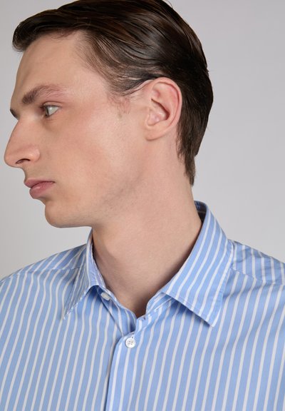Młody mężczyzna z zaczesanymi do tyłu brązowymi włosami, ubrany w niebiesko-białą koszulę w pionowe paski, zwrócony w lewo na jednolitym tle.