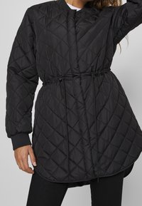 Svart vadderad lång jacka med åtdraget midja, ribbade manschetter och dragsnörning. Har ett slätt, texturerat tyg med diamantmönster.