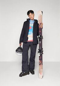 Άτομο σε μαύρη στολή σκι κρατώντας ένα κράνος και ένα ζευγάρι σκι σε όρθια θέση, φορώντας γυαλιά σκι και χειμερινά μποτίνια σε απλό φόντο.