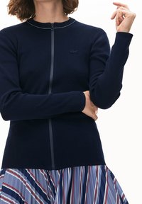 Mörkblå zip-up kofta med rund halsringning, prydd med en tunn silverdetalj och Lacoste-logotyp. Matchas med en veckad randig kjol.