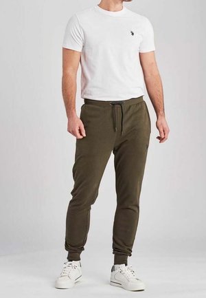 Mann steht und trägt ein weißes Kurzarmhemd, olivgrüne Jogginghosen mit Kordelzug und weiße Sneakers auf grauem Hintergrund.