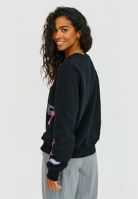 Čierna mikina s výraznými grafickými potlačami na rukávoch. Má okrúhly výstrih a rebrované manžety. Nosená s svetlosivými pruhovanými nohavicami.