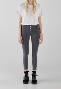 Mujer rubia que lleva una camiseta blanca de manga corta, unos jeans skinny de cintura alta en gris oscuro con botones y botas negras con cordones.