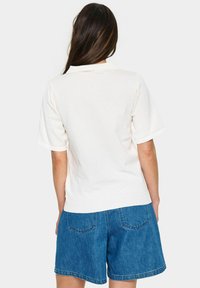 Weißer Kurzarmpullover mit glattem Strickmuster und geripptem Saum, kombiniert mit blauen Jeansshorts mit zwei Gesäßtaschen.