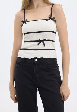 Femme portant un haut sans manches blanc texturé avec des finitions noires et des nœuds, associé à un jean noir taille haute, debout devant un fond uni.