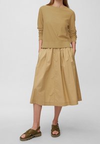 Camicia kaki a maniche lunghe abbinata a una gonna beige plissettata al ginocchio e sandali verde oliva con design a fascia incrociata.