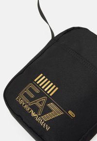Borsa di tessuto nero con logo dorato "EA7 EMPORIO ARMANI" e strisce orizzontali, texture liscia e chiusura con zip.
