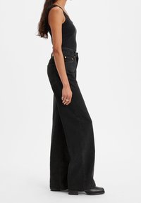 Jean en denim noir à taille haute et jambe large avec cinq poches. Présente une coupe droite et un ourlet effiloché. Assorti à un débardeur noir ajusté.