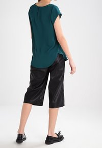 Teal blouse met korte mouwen, een ronde hals en een sleutelgatdetail aan de achterkant, gecombineerd met zwarte, cropped, glanzende broeken en zwarte platte schoenen.