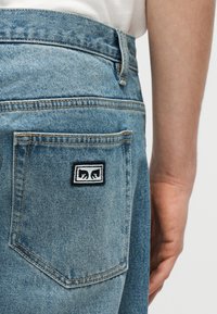 Artimas šviesiai mėlynos spalvos džinsinių kelnių galinės kišenės vaizdas, ant kurios yra mažas stačiakampis juodas ir baltas pleistras su dviem juodomis rankų piktogramomis.