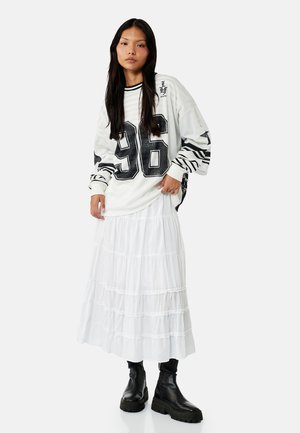 Kobieta o długich ciemnych włosach, ubrana w oversize’owy biały sweter z napisem „96”, białą warstwową spódnicę i czarne masywne buty, stoi na tle jednolitego tła.