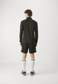 Schwarzes langärmliges Oberteil, schwarze Shorts und knielange weiße Socken mit schwarzen Streifen. Die Person trägt schwarze Sportschuhe.
