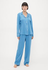 Μπλε πιτζάμα από μαλακό ύφασμα. Μπλουζάκι με μακριά μανίκια και κουμπιά στο μπροστινό μέρος με λευκή περιθώριο, σε συνδυασμό με φαρδιά παντελόνια. Χωρίς σχέδια.