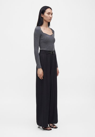 Femme aux longs cheveux noirs portant un haut gris ajusté à manches longues, un pantalon large noir taille haute, une ceinture noire et des sandales noires à talons.