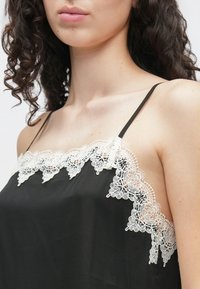 Czarna satynowa koszulka na cienkich, regulowanych ramiączkach z misterną, białą koronką wzdłuż dekoltu i gładką fakturą.
