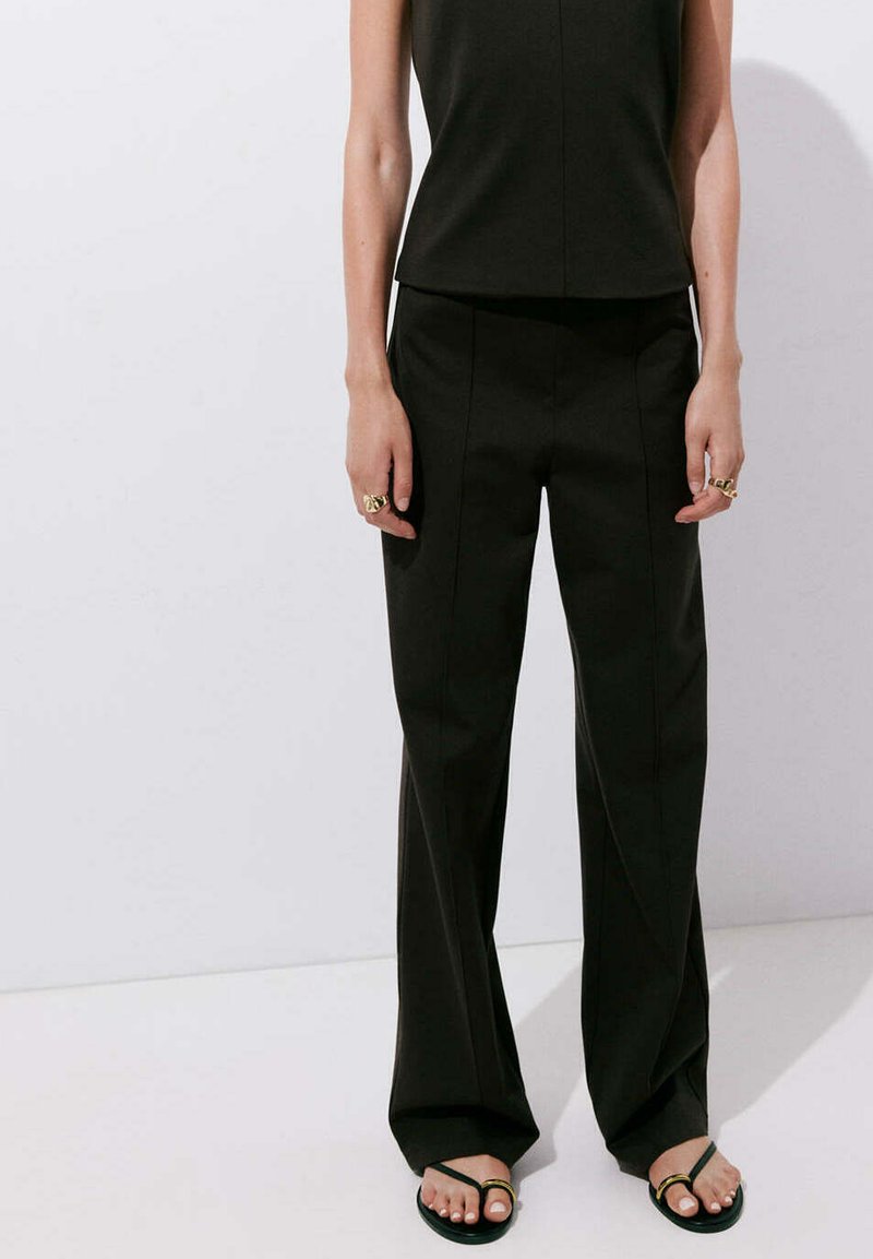 Personne portant un haut noir sans manches, un pantalon noir à jambes larges, des bagues dorées et des sandales noires avec des accents dorés, debout contre un mur blanc.