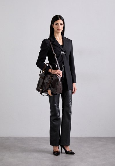 Schwarzer Blazer mit einem silbernen Verschluss, kombiniert mit schwarzen Lederhosen und schwarzen Absatzschuhen. Eine braune Lederhandtasche mit Schultergurt rundet den Look ab.