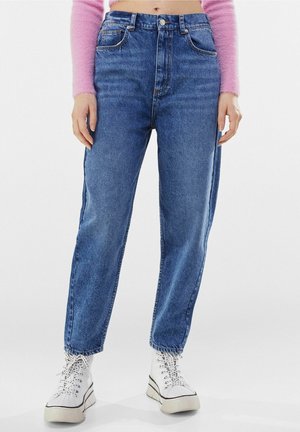 Personne portant un jean bleu taille haute, des baskets à plateforme blanches avec lacets, et un pull court en laine rose à manches longues, debout sur un sol blanc.