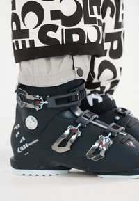Zwarte skschoenen met metalen gespen, met een gladde afwerking en gestructureerde accenten. Grijze broekspijpen met vetgedrukte zwarte letters erboven.