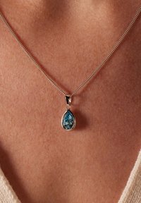 Collier en chaîne en argent avec un pendentif en forme de goutte d'eau en pierre précieuse bleue, à facettes claires et à finition polie, reposant contre la peau.