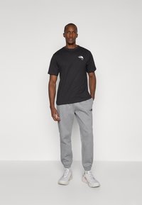 T-shirt en coton noir avec un petit logo sur la poitrine, associé à un pantalon de jogging gris en tissu doux. Modèle portant des baskets blanches.