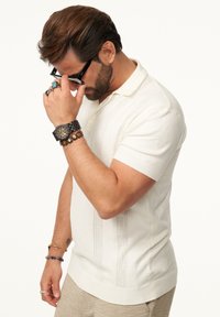 Weißes Poloshirt mit kurzen Ärmeln, einem strukturierten Design und vertikalen Streifen im Akkordeon-Stil. Getragen mit einer Uhr und mehreren Armbändern.
