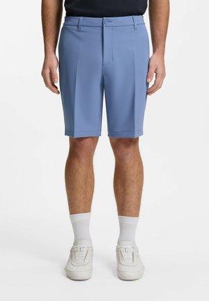 Uomo che indossa pantaloncini azzurri su misura, calzini bianchi e scarpe da ginnastica bianche, in piedi contro uno sfondo bianco uniforme.