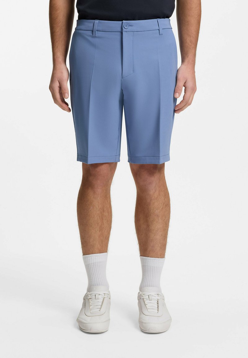 Homme portant un short bleu clair sur mesure, des chaussettes blanches et des baskets blanches, debout devant un fond blanc uni.