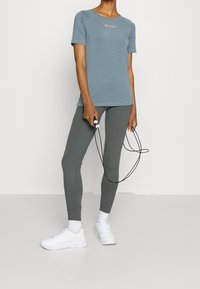 T-shirt de sport à manches courtes bleu clair avec logo orange, associé à des leggings gris et des baskets blanches, tenant une corde à sauter.