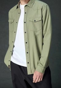 Camicia a maniche lunghe di un verde oliva chiaro con due tasche sul petto, bottoni neri e una consistenza morbida, indossata sopra una maglietta bianca e pantaloni neri.