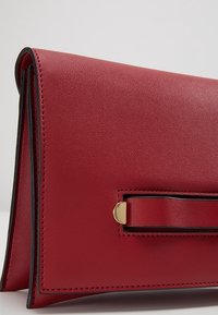 Rode leren clutch met een gladde textuur, voorzien van goudkleurige hardware-accenten en een slank, gestructureerd ontwerp. Vouwranddetail zichtbaar.