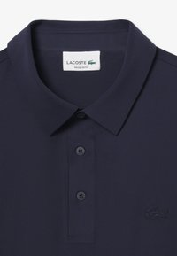 Polo Lacoste azul marino con cuello clásico, dos botones y logo de cocodrilo bordado en el pecho, etiquetado como ajuste regular.
