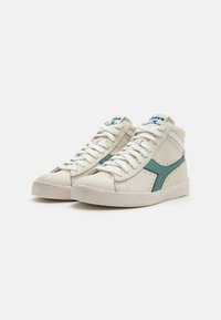 Sneakers alte in pelle bianca sporca con dettagli teal, dotate di lacci bianchi, punte perforate e suola in gomma testurizzata.