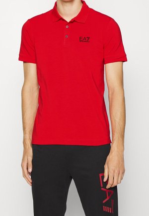 Homme portant un polo rouge avec le logo noir EA7 Emporio Armani et un pantalon noir avec l'inscription rouge EA7 sur la cuisse.