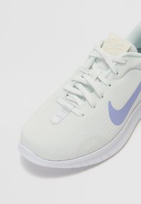 Vaaleansininen urheilusandaali, jossa on teksturoidusta kankaasta valmistettu päällinen, valkoiset nauhat ja violetti Nike-logon swoosh. Mukana pyöreä kärki ja sileä pohja.