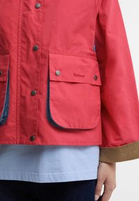 Jachetă roșie cu buzunare mari în față, cu închideri cu capse, tiv maro la mâneci, purtată peste o cămașă albastru deschis și pantaloni închiși la culoare pe o persoană în picioare.