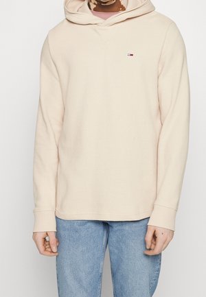 Osoba w beżowym, teksturowanym hoodie z małym logo na piersi oraz jasnoniebieskich dżinsach, stojąca na jednolitym tle.