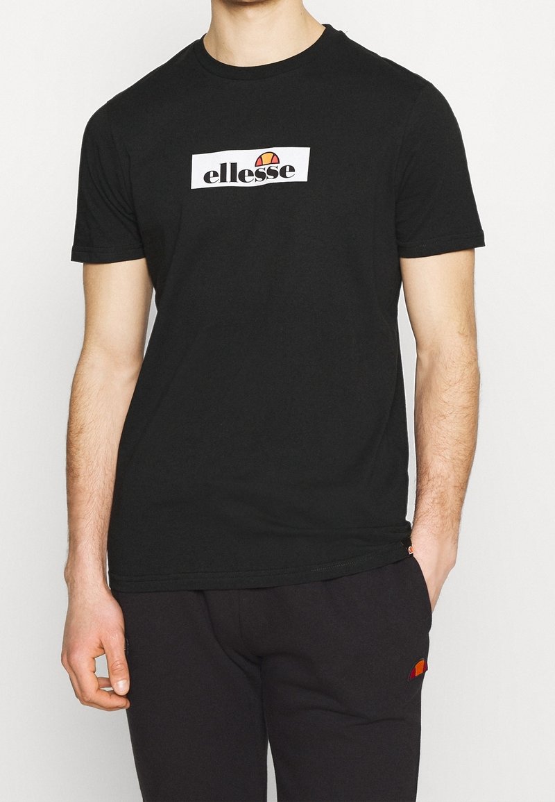 Homme portant un t-shirt noir à manches courtes avec le logo blanc "ellesse" et un pantalon noir assorti avec un petit logo orange sur la poche.