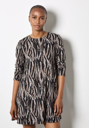 Frau mit kurzen Haaren, die ein langärmliges, zebra gestreiftes Kleid trägt und vor einer weißen Wand steht.