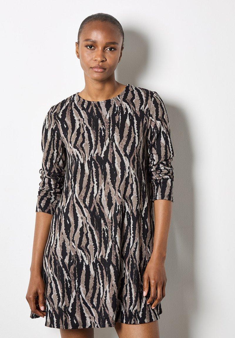 Frau mit kurzen Haaren, die ein langärmliges, zebra gestreiftes Kleid trägt und vor einer weißen Wand steht.