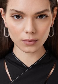 Silver hoop earrings with a rectangular shape, featuring intricate details and small stones along the edge, worn by a model.