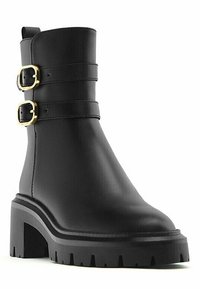 Bottine en cuir noir avec un talon épais et une semelle crantée, dotée de deux sangles dorées à boucle autour de la tige supérieure.