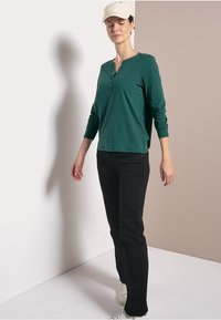 Zelená dlouhá halenka s knoflíky, černé kalhoty a bílé tenisky. Model nosí krémovou čepici a pózuje na neutrálním pozadí.