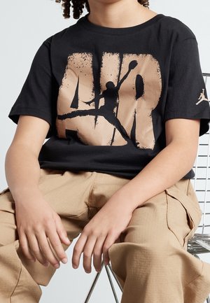 Enfant portant un t-shirt noir avec de grandes lettres beige "AIR" et la silhouette d'un joueur de basket, assis avec un pantalon cargo beige sur une chaise en métal.