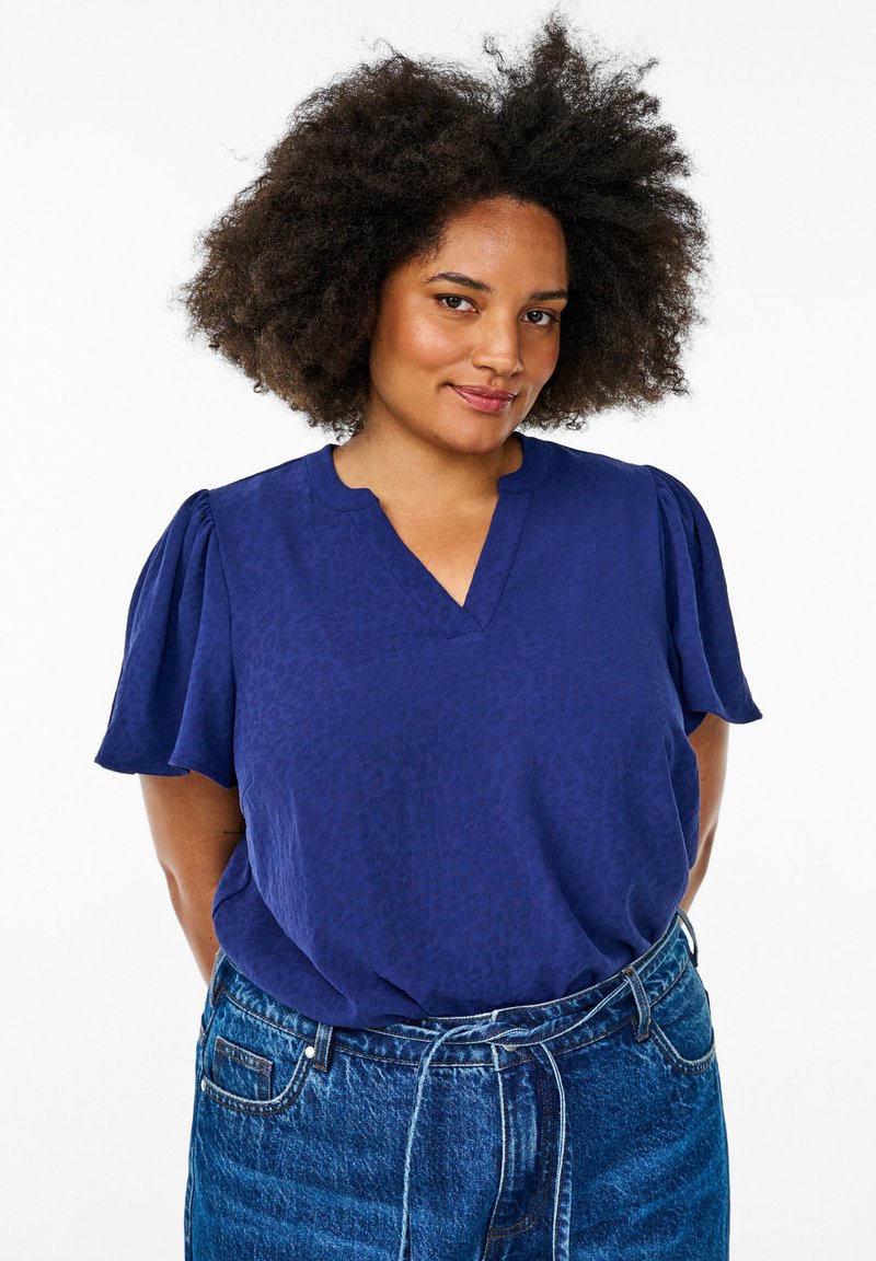 Blouse courte bleu marine à manches courtes avec un col en V et un motif texturé de léopard ; portée avec un jean en denim taille haute avec une ceinture à nouer.
