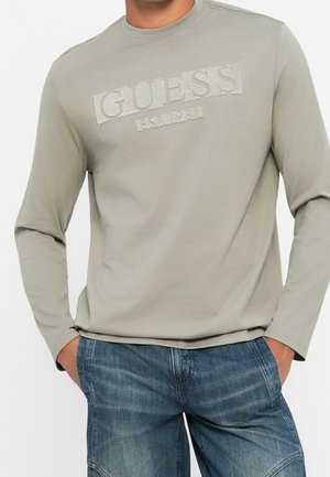 Mężczyzna w jasno szarej koszuli z długim rękawem z teksturowanym logo "GUESS" i niebieskich jeansach, ręce w kieszeniach.
