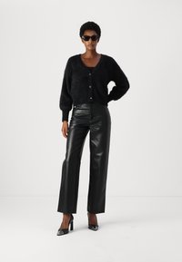 Cardigan nero peloso con scollatura a V, chiusura con bottoni, abbinato a pantaloni in pelle nera a gamba larga e scarpe con tacco alto con un accento lucido sulle punte.